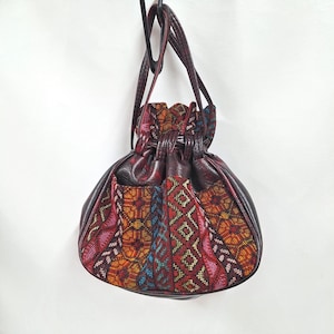 May include: A round, burgundy drawstring bag with a patterned fabric panel featuring red, orange, and blue geometric designs. The bag has a leather-like texture and a long strap for carrying.