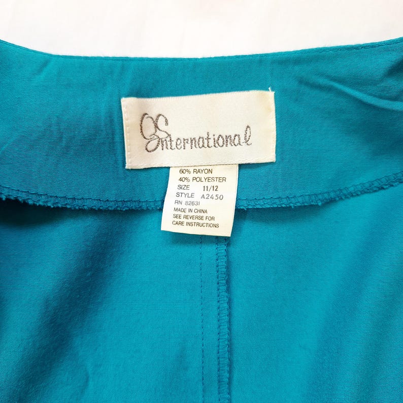 May include: A teal garment with a white label showing the brand "International". The label details the fabric composition, size 11/12, style A2450, and RN 82631. The garment is made in China.