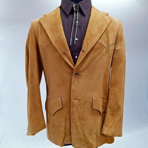 May include: A tan suede blazer featuring a notched lapel, two front flap pockets, and a single button closure. The blazer is worn over a dark brown button-down shirt with a silver-toned bolo tie featuring a decorative stone centerpiece.