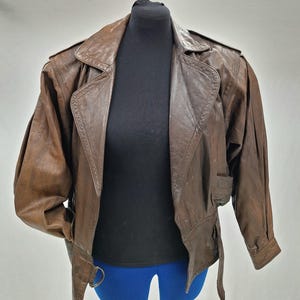 May include: A brown leather jacket with a wide collar, shoulder pads, and a belt. The jacket has long sleeves and a tailored fit. The jacket is displayed on a black mannequin.