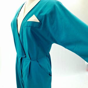 May include: Teal jumpsuit with a V-neck and long sleeves. The jumpsuit features a cream-coloured lapel and a pocket with a cream-coloured handkerchief. The garment has a relaxed fit and a button closure at the waist.