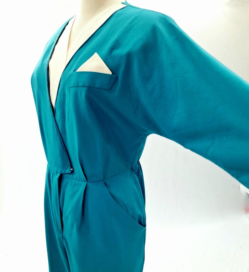 May include: Teal jumpsuit with a V-neck and long sleeves. The jumpsuit features a cream-coloured lapel and a pocket with a cream-coloured handkerchief. The garment has a relaxed fit and a button closure at the waist.