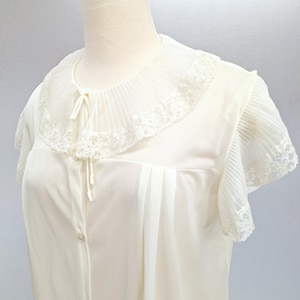 May include: A cream-colored short-sleeve blouse featuring a pleated round collar with white lace trim. The garment includes a front button closure with a small tie detail at the neckline and pleated sleeve cuffs finished with matching lace edging.