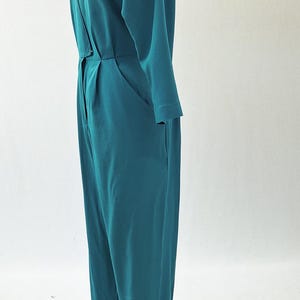 May include: Teal jumpsuit with long sleeves and a V-neckline. The one-piece garment features a button closure at the waist, pockets, and a small pocket on the chest. The jumpsuit is displayed on a mannequin.