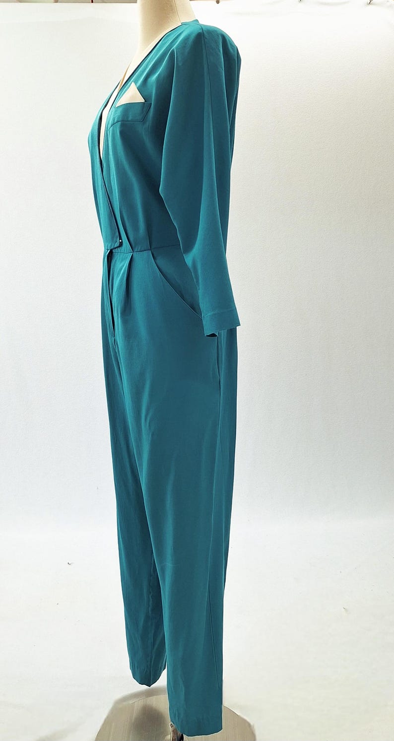 May include: Teal jumpsuit with long sleeves and a V-neckline. The one-piece garment features a button closure at the waist, pockets, and a small pocket on the chest. The jumpsuit is displayed on a mannequin.