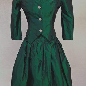 May include: A dark emerald green two-piece dress set featuring a fitted jacket with an off-the-shoulder neckline, three-quarter length sleeves, and three circular silver-toned buttons. The matching skirt has a gathered, pleated waistline.