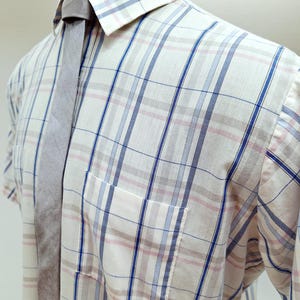May include: A short-sleeved, button-down shirt with a plaid pattern in white, blue, and pink. A gray tie is knotted at the collar. The shirt has a chest pocket and is displayed on a mannequin.