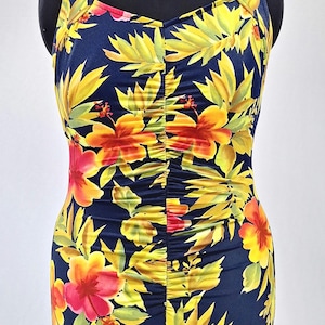 May include: A one-piece swimsuit with a vibrant floral pattern. The swimsuit features a dark blue background adorned with yellow, orange, and red tropical flowers and green leaves. The fabric is gathered down the center. The swimsuit is displayed on a black mannequin.