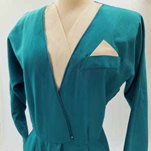May include: Teal jumpsuit with long sleeves and a V-neck. It has a cream-coloured lapel and pocket square. The design includes a front pocket and a tailored fit, showcasing a vintage style. The jumpsuit is a classic piece.