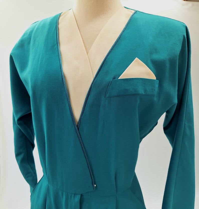 May include: Teal jumpsuit with long sleeves and a V-neck. It has a cream-coloured lapel and pocket square. The design includes a front pocket and a tailored fit, showcasing a vintage style. The jumpsuit is a classic piece.