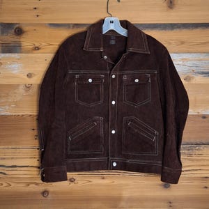 May include: A brown suede jacket with white stitching and four front pockets. The jacket has a button-up closure and a collar. It is hanging on a white hanger against a wooden background.