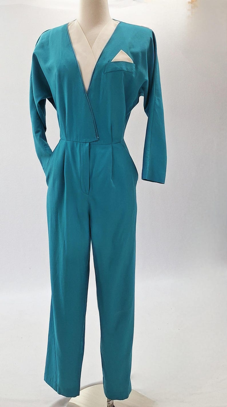May include: Teal jumpsuit with a V-neck and long sleeves. It features a cream-coloured lapel and pocket detail. The waist is cinched, and the trousers have a wide-leg design. The garment is displayed on a mannequin.