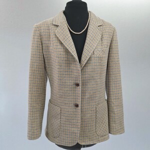 May include: A tan and blue plaid blazer featuring a notched lapel, two brown buttons for closure, a single chest pocket, and two lower patch pockets. The blazer is displayed on a black mannequin wearing a simple white beaded necklace.