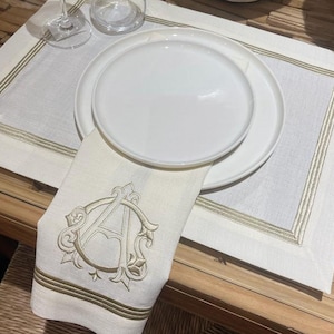 Può includere: Un tovaglietta di lino bianco con un bordo ricamato in oro. Un piatto bianco con un tovagliolo ricamato in oro è posto sopra la tovaglietta.