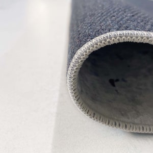 Puede incluir: Primer plano del borde de una alfombra gris oscuro con un borde blanco cosido.