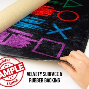 May include: A black rug with a colorful design featuring the PlayStation symbols. The rug has a velvety surface and a rubber backing. The text "SAMPLE" is printed on a red circle.