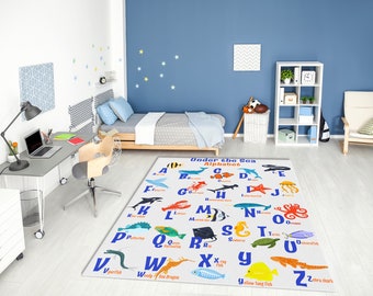 May include: A white rug with a colorful alphabet printed on it. Each letter is paired with a sea creature, such as a fish, crab, or octopus. The rug is in a child's bedroom with a bed, desk, and bookshelf in the background.