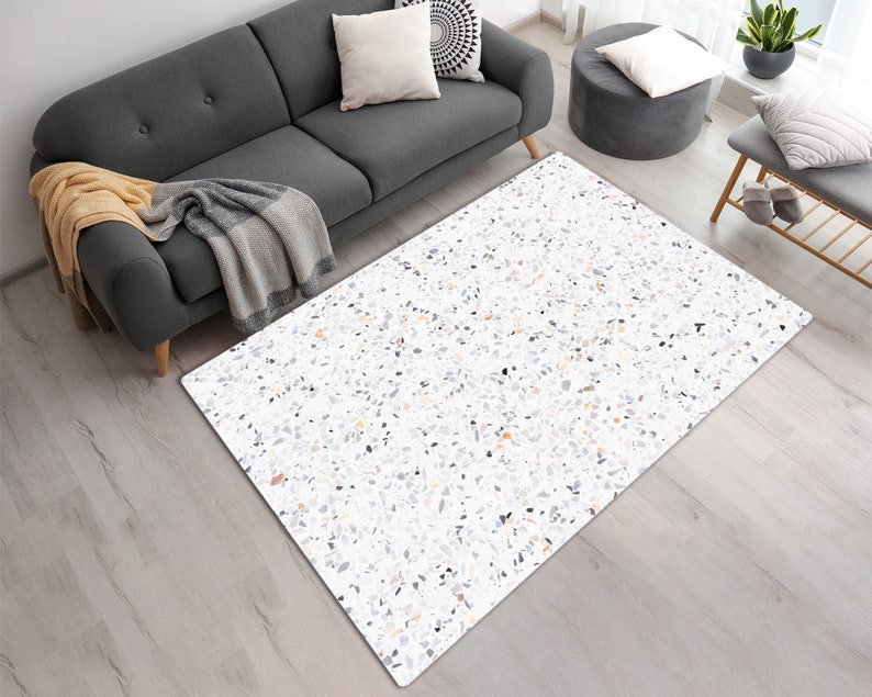 Puede incluir: Una alfombra de estilo terrazo blanco y gris con un fondo gris claro y peque&ntilde;as motas negras, blancas y grises. La alfombra est&aacute; sobre un suelo de madera clara.