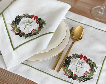 May include: White linen napkin with a green border and embroidered Christmas wreath design. The wreath features red berries, green leaves, and a red bow. A matching placemat and gold utensils are also visible.