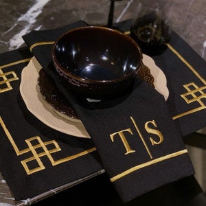 Könnte beinhalten: Schwarze Tischsets mit goldenen geometrischen Mustern und eine schwarze Serviette mit goldener Stickerei, die "T|S" lautet.