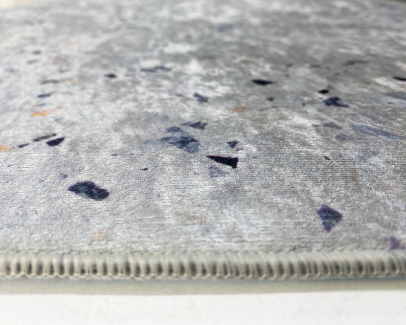 Puede incluir: Primer plano de una alfombra gris con un sutil patr&oacute;n moteado. La alfombra tiene un borde cosido.