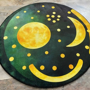 Pode incluir: Um tapete redondo com fundo verde escuro e uma lua amarela, uma lua crescente e outras formas. O tapete tem um brilho sutil.