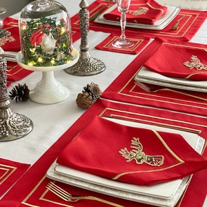 Puede incluir: Manteles individuales rojos con ribete dorado y diseño de reno. Los manteles individuales están colocados sobre un mantel blanco con candelabros de plata y una campana de cristal con un pequeño árbol de Navidad en su interior.