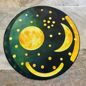 Pode incluir: Um tapete redondo verde e amarelo com um desenho de lua crescente e sol. O tapete tem um fundo verde escuro com pontos amarelos e uma lua crescente e um sol amarelos.