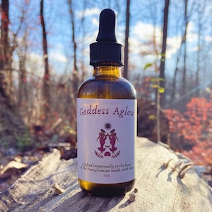 May include: A brown glass bottle with a dropper top, labeled "Goddess Aglow." The label features a design of two figures and text. The bottle is 2 oz. and is set against a blurred forest background.