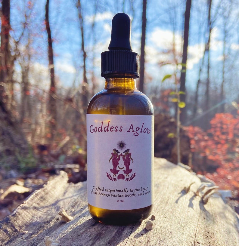 May include: A brown glass bottle with a dropper top, labeled "Goddess Aglow." The label features a design of two figures and text. The bottle is 2 oz. and is set against a blurred forest background.
