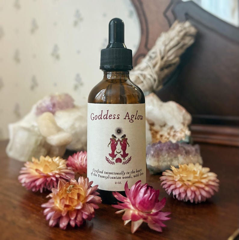 May include: A brown glass bottle with a black dropper, labeled "Goddess Aglow." The label features a design of two figures and text. The bottle is surrounded by dried flowers in shades of pink and orange, with crystals in the background.