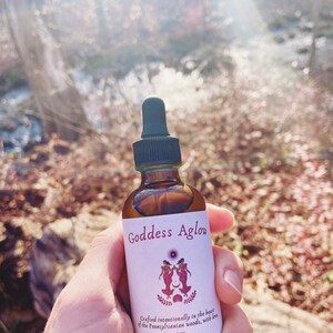 May include: A brown glass bottle with a black dropper, labeled "Goddess Aglow." The label features two figures, a sun, and the text "Crafted intentionally in the heart of the Pennsylvanian woods, with love." The bottle contains 2 oz of liquid.