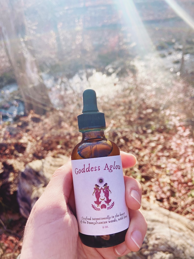 May include: A brown glass bottle with a black dropper, labeled "Goddess Aglow." The label features two figures, a sun, and the text "Crafted intentionally in the heart of the Pennsylvanian woods, with love." The bottle contains 2 oz of liquid.