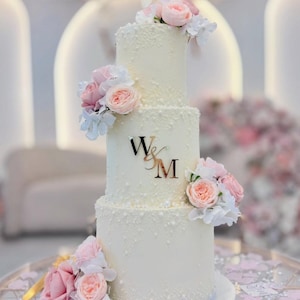 May include: A three-tiered white wedding cake decorated with pink and white flowers. The cake has a textured surface and features the initials "W&M" in gold. The cake is on a glass table.