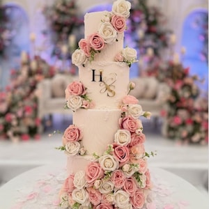 May include: A four-tiered wedding cake decorated with pink and white roses. The cake has a light pink frosting and features the initials "H & S" in gold. Pink and white rose petals are scattered around the base.