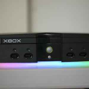 May include: Black Xbox console with the word "XBOX" in white lettering. The console has a multi-colored light strip along the bottom. The console has two sets of ports on the front.