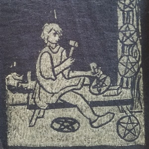 Peut inclure: Un écusson en tissu bleu foncé avec une illustration claire d'une figure travaillant avec des outils. Le motif comprend un pentagramme et d'autres symboles, avec une structure en forme de colonne sur le côté droit.