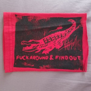 May include: A red and black textile item featuring a red alligator illustration and the text "FUCK AROUND & FIND OUT". The design is printed on a rectangular piece of fabric.