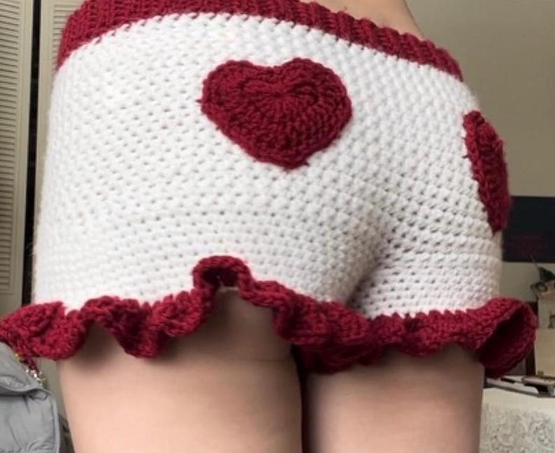 K&ouml;nnte beinhalten: Wei&szlig;e H&auml;kelshorts mit roter Herzapplikation und roter Bord&uuml;re. Die Shorts haben einen ger&uuml;schten Saum und ein hoch tailliertes Design. Die H&auml;kelarbeit ist detailliert und die Farbkombination ist auff&auml;llig.