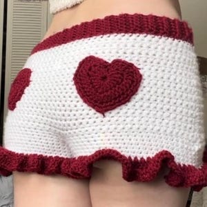 K&ouml;nnte beinhalten: Wei&szlig; geh&auml;kelte Shorts mit rotem Herzdesign auf der Vorder- und R&uuml;ckseite. Die Shorts haben einen roten R&uuml;schenbesatz am Saum und ein rotes Band am Bund. Handgefertigt.
