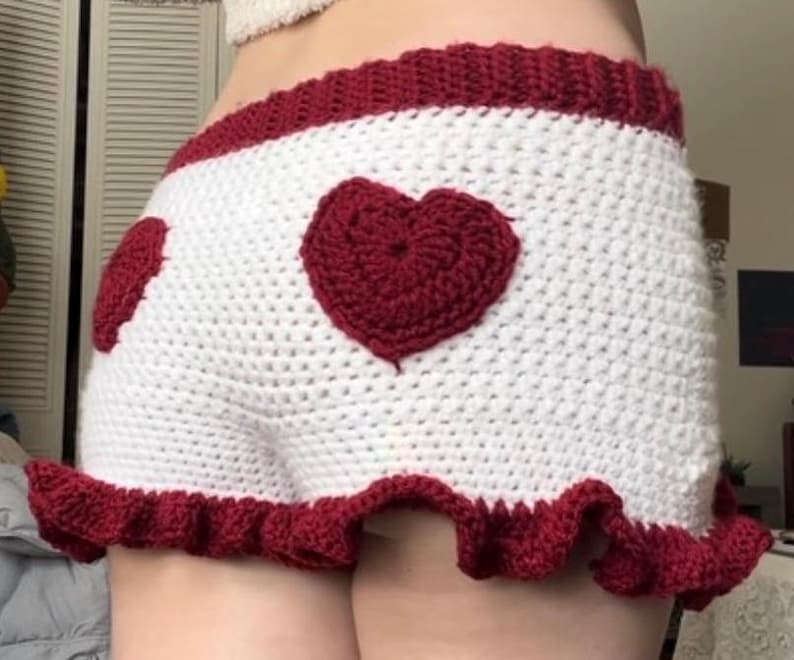 K&ouml;nnte beinhalten: Wei&szlig; geh&auml;kelte Shorts mit rotem Herzdesign auf der Vorder- und R&uuml;ckseite. Die Shorts haben einen roten R&uuml;schenbesatz am Saum und ein rotes Band am Bund. Handgefertigt.