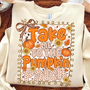 May include: Cream-colored sweatshirt with the text "Take Me To The Pumpkin Patch" in orange and peach lettering. The design includes pumpkins, sunflowers, and fall leaves. A brown bow is at the top left of the design.