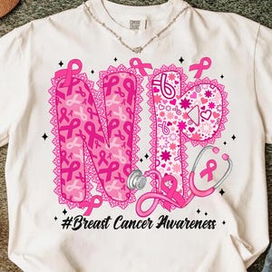 May include: Cream-colored t-shirt with a pink design promoting breast cancer awareness. The design features the letters "NP" filled with pink ribbons, a stethoscope, and the text "#Breast Cancer Awareness."