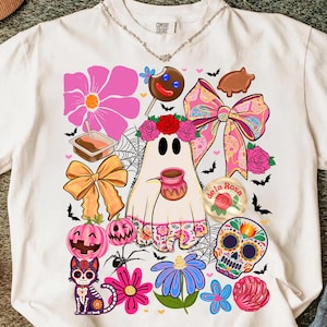 May include: White t-shirt with a colorful Halloween-themed graphic. The design includes a ghost holding a mug, a sugar skull, pumpkins, flowers, bows, and a cat. The shirt also features the text "de la Rosa".