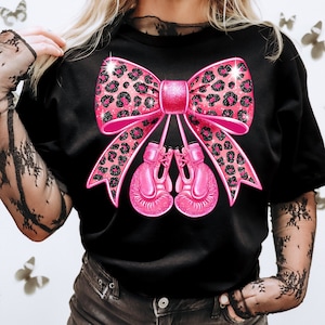 May include: Black t-shirt with a pink leopard print bow and pink boxing gloves. The bow has glitter accents.