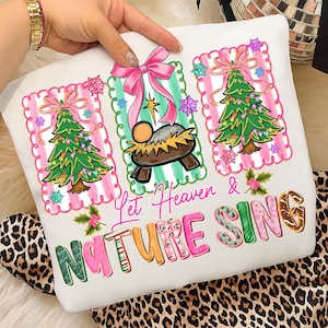 May include: White Christmas-themed pillow with three framed illustrations of a Christmas tree, a manger scene, and the text "Let Heaven & Nature Sing." The illustrations are framed with pink and green borders and adorned with pink bows and snowflakes.