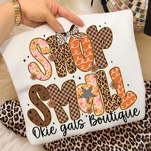 May include: White t-shirt with the words "Shop Small" in a colorful, patterned design. The letters are decorated with floral, leopard print, and checkered patterns. The text "Okie gals Boutique" is written below the main text.