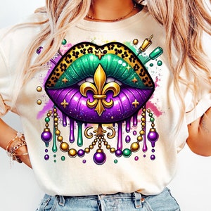 May include: A cream-coloured t-shirt with a detailed graphic of stylised lips. The lips are a mix of purple, green, and gold, with a leopard print upper lip and a gold fleur-de-lis. Beaded accents and paint splatters complete the design.