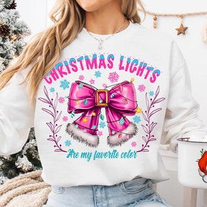 以下が含まれることがあります： クリスマスライトが大好きな色という文字が入った、ピンクのリボンとクリスマスライトのデザインが施された白いスウェットシャツ。