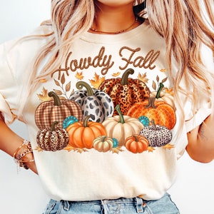 May include: A beige t-shirt with the text "Howdy Fall" in a rope-like font. The shirt features a cluster of colorful pumpkins with various patterns, including plaid, leopard print, and turquoise accents.